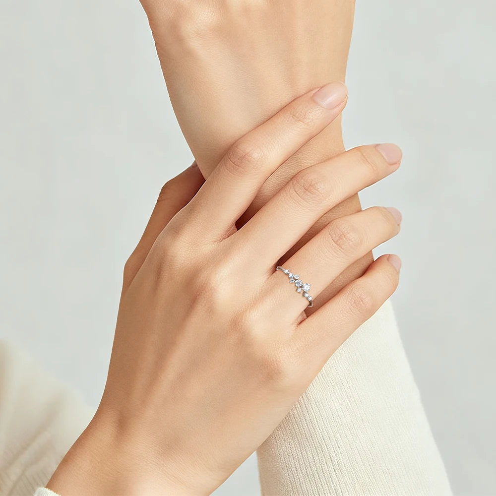 A delicate silver ring with small clear stones worn on a woman's ring finger, shown on her hand against a soft light gray background.