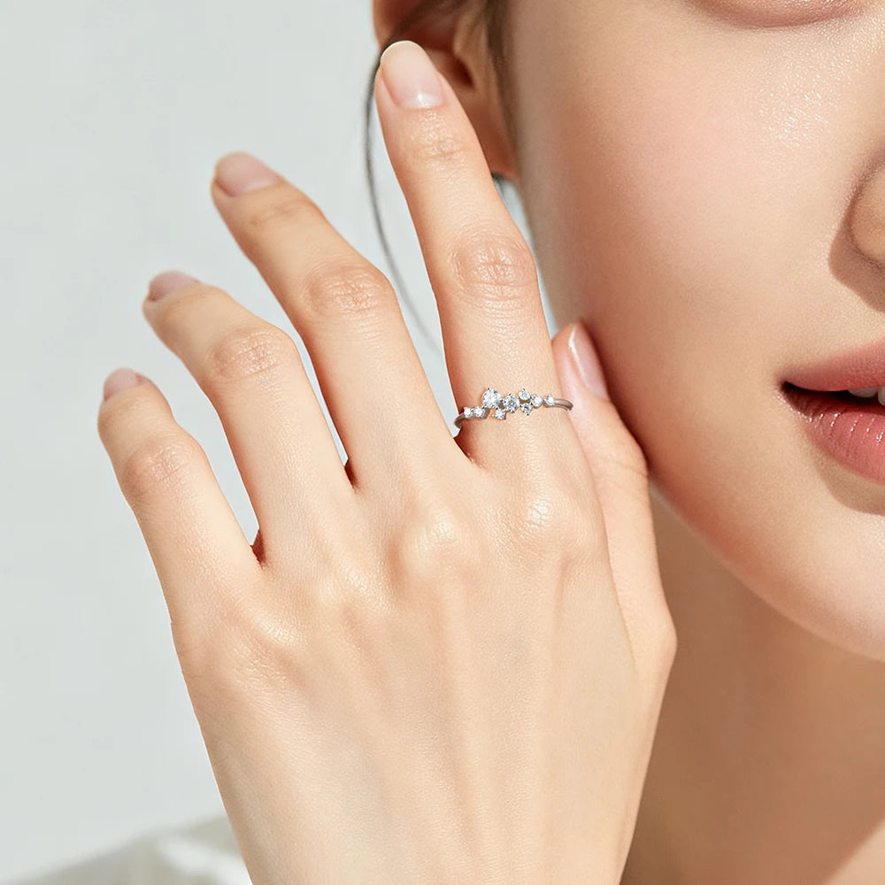 A close-up lifestyle shot of a model's hand wearing a delicate silver ring adorned with small clear gemstones, set against a soft, light gray background.