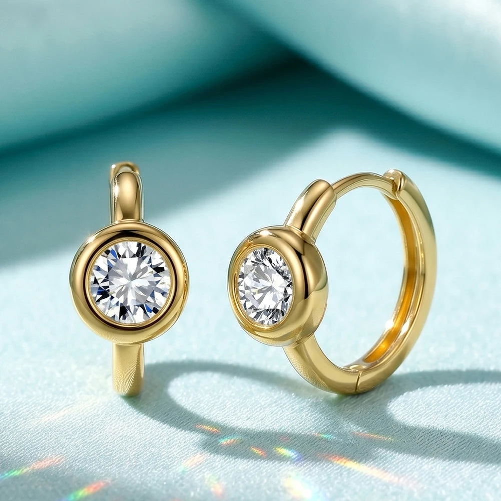 A pair of gold hoop earrings with a round bezel-set clear gemstone, displayed on a light blue textured fabric background.