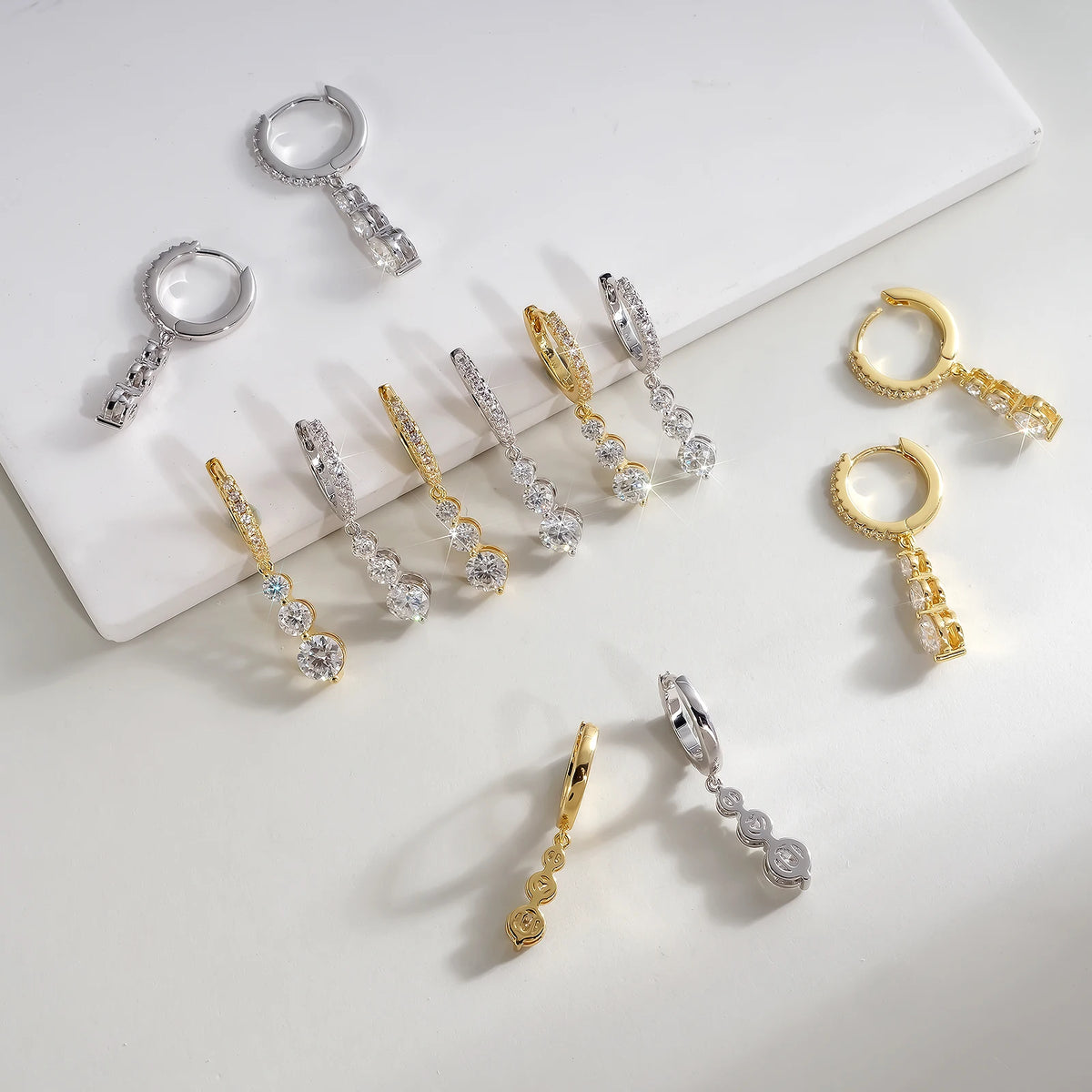 Nine pairs of huggie hoop earrings with dangling crystal bead chains are arranged on a white surface and a white rectangular tray. The earrings are shown in silver, yellow gold, and two-tone silver and gold finishes.