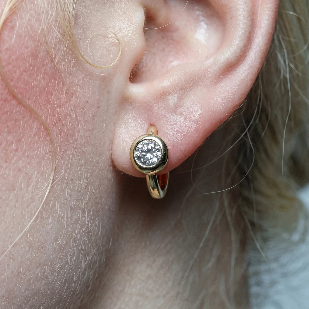 A close-up of a person's ear wearing a small gold huggie hoop earring with a round clear gemstone in a bezel setting.