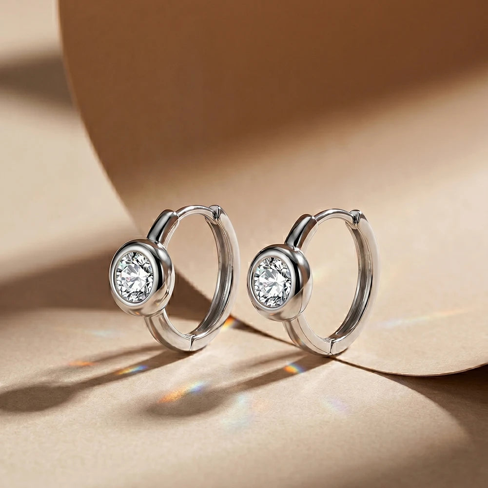 A pair of silver hoop earrings with a round bezel-set clear gemstone, displayed on a warm beige textured surface with soft shadows.