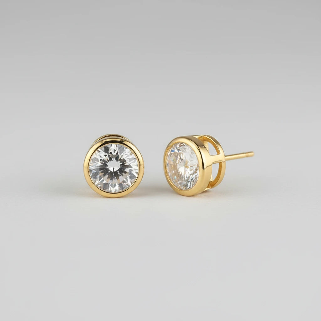 A pair of gold stud earrings with oval-cut clear gemstones, shown on a warm beige surface with soft shadows.