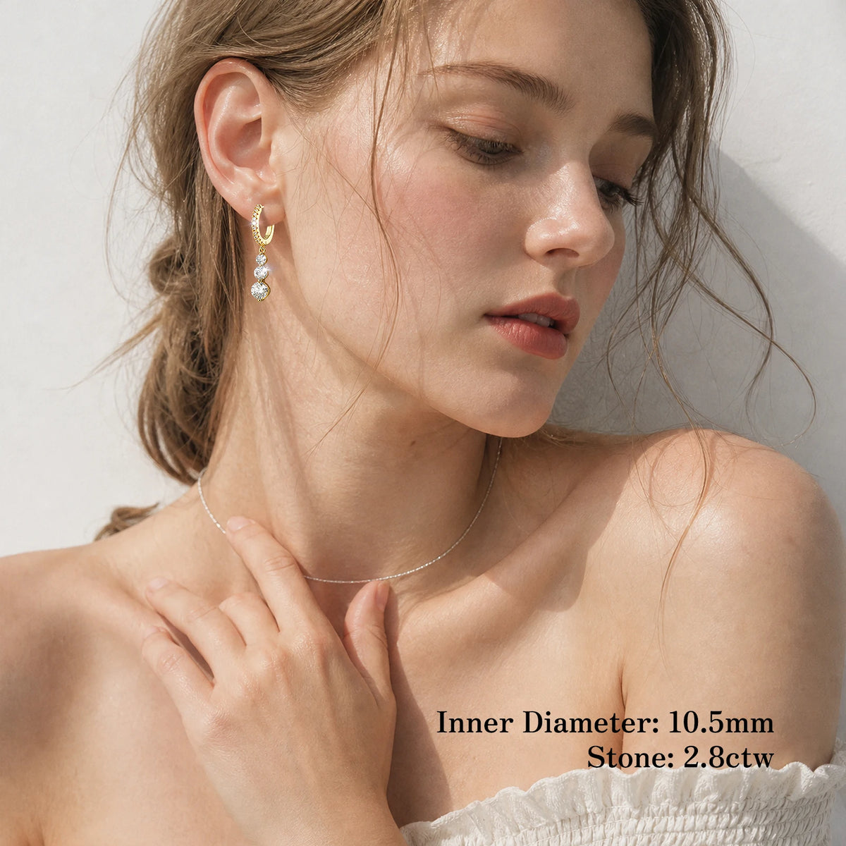 A woman wearing delicate jewelry, including a thin silver chain necklace and a gold hoop earring with clear stones, shown against a light wall with text overlay detailing the earring's dimensions.