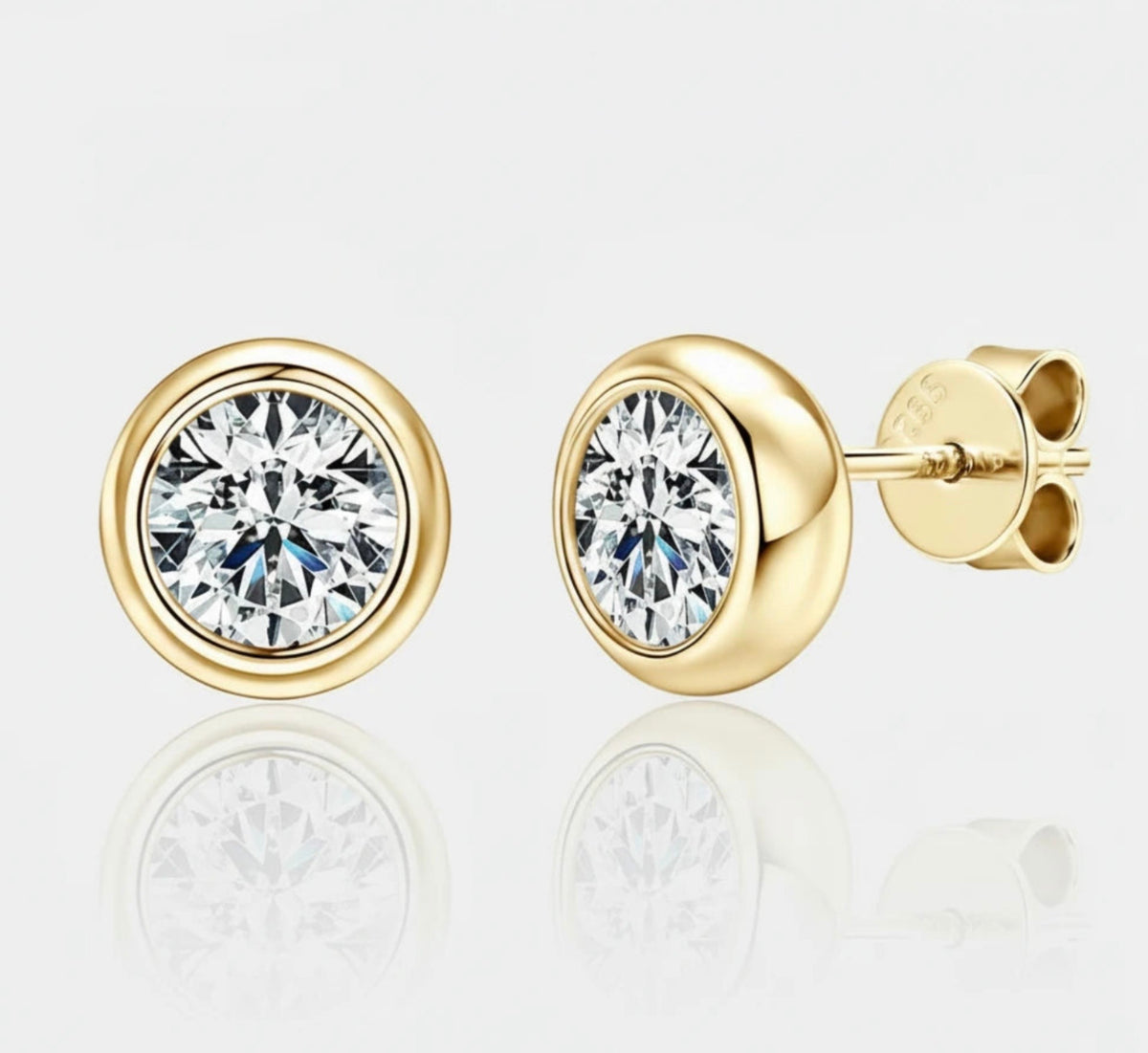 A pair of gold stud earrings with round bezel-set clear gemstones, shown on a light gray reflective background. One earring is facing forward, and the other is turned to show the post and backing.