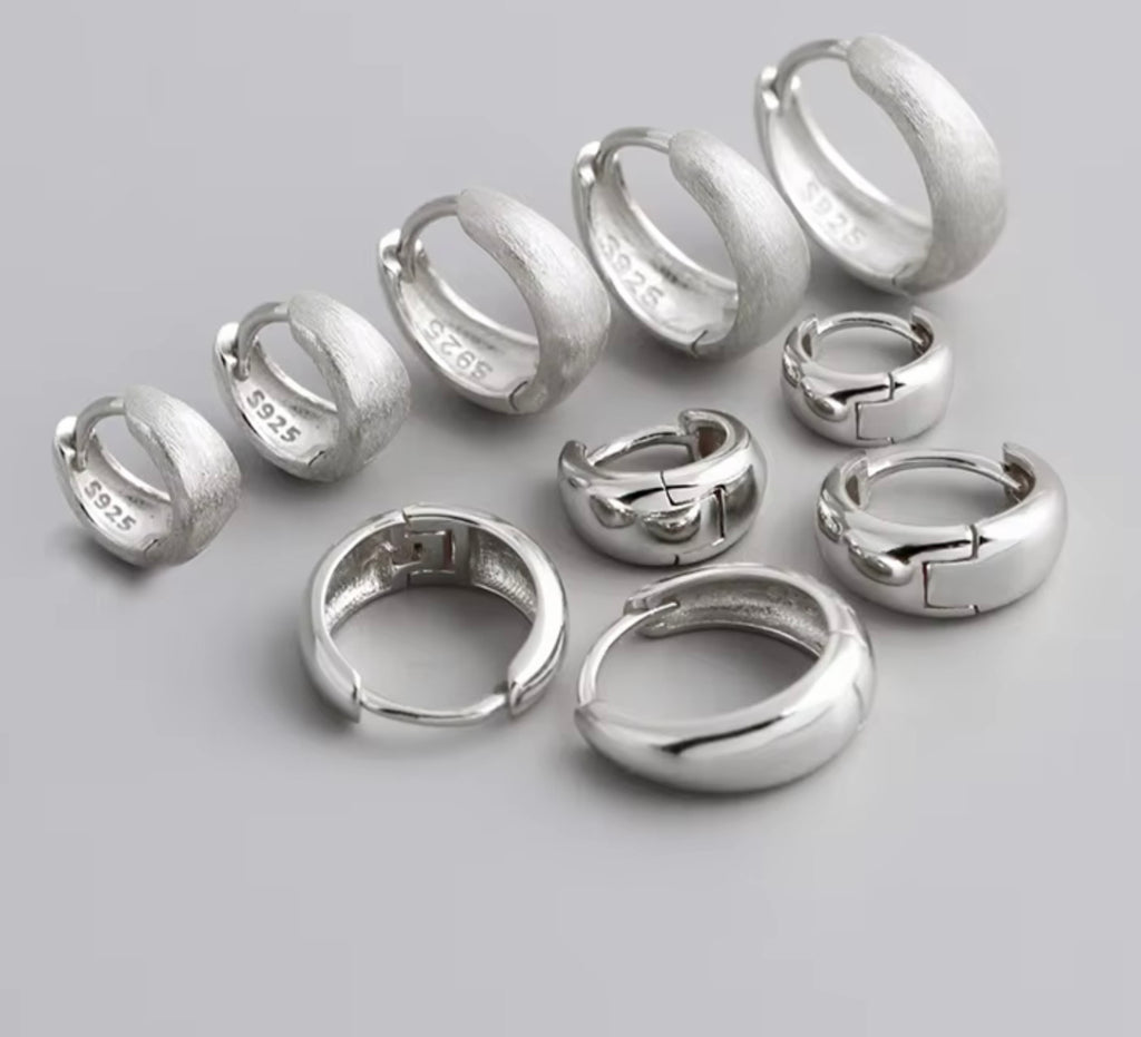 A set of silver huggie hoop earrings in various sizes, displayed on a neutral gray background. Several earrings show the sterling silver purity stamp '925' on the inside.