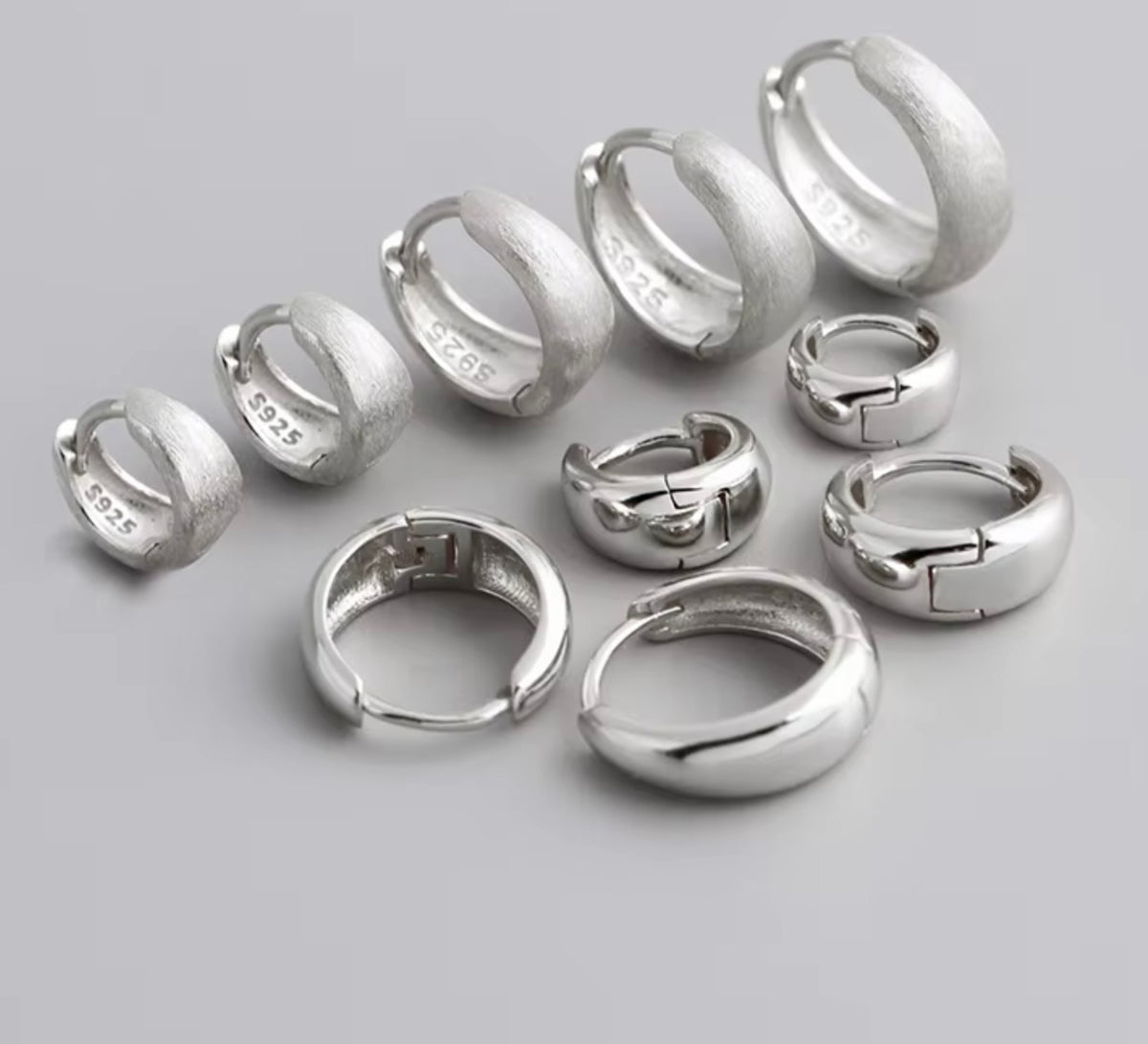 A set of silver huggie hoop earrings in various sizes, displayed on a neutral gray background. Several earrings show the sterling silver purity stamp '925' on the inside.